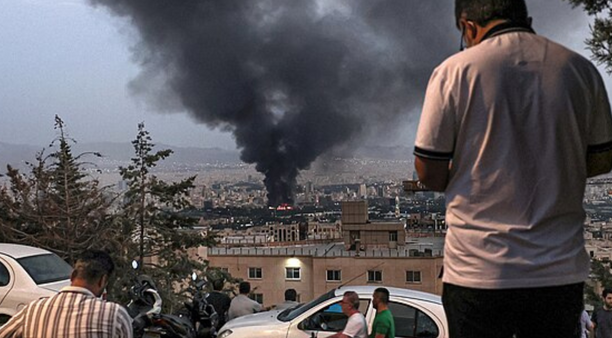 Two men on a hill look down on a building hit by Israeli airstrikes in Tehran. In front of them, two men pass on a motorbike.