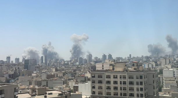 A city scape of Tehran, showing various buildings in the background on fire from Israeli bombs.