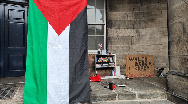 student encampment at the University of Edinburgh, a Palestinian flag stands before a cardboard sign saying "Walid Daqqa Library"