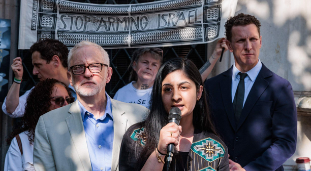 Zarah Sultana holds a microphone in front of Jeremy Corbyn at a rally. Behind them, two people hold a sign saying "Stop Arming Israel"