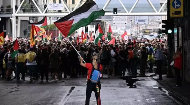 A woman waving a Palestinian flag stands with her back to the camera. A large crowd amasses in the background.