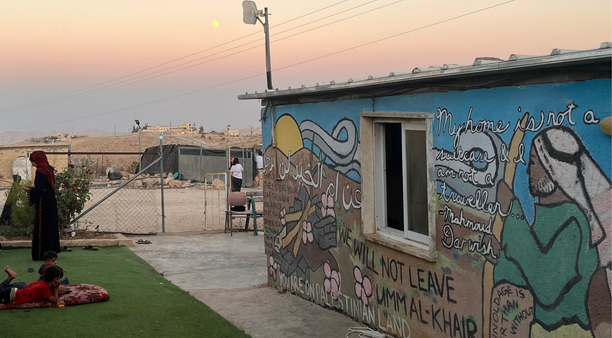 An image of a mural in the Palestinian village of Umm al Khair. The mural shoes a Shepard in a Keffiyah on the hills of the village, with a Mahmoud Darwish quote and the declaration "we will not leave Umm al-Khair"