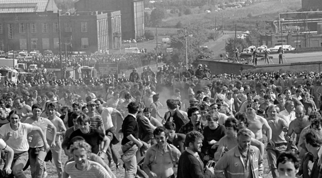 Black and white image of a group of miners running from the police at Orgreave, 1984.