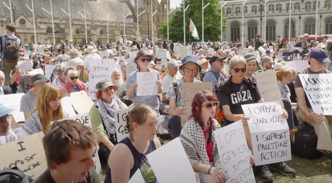 A large group of demonstrators sit in Parliament Square, London, holding placards that say "I oppose genocide, I support Palestine Action"