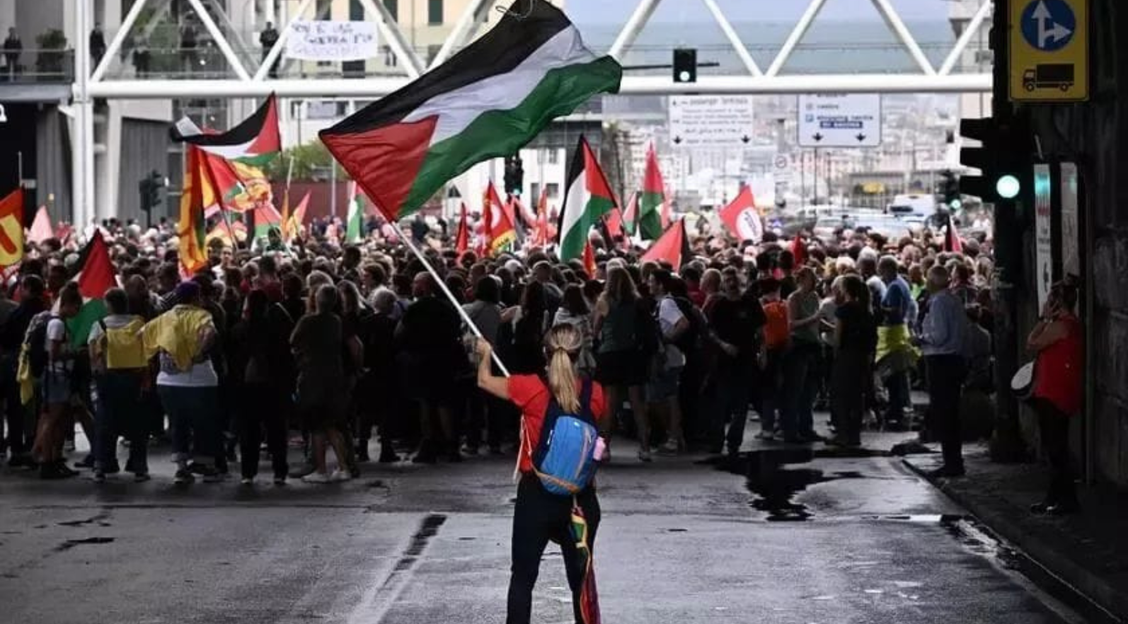 A woman waving a Palestinian flag stands with her back to the camera. A large crowd amasses in the background.